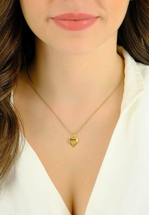 Collier avec pendentif en or en forme de cœur sur une fine chaîne, présentant un design tordu. Le pendentif est petit et brillant, reposant au-dessus d'une chemise blanche à col.