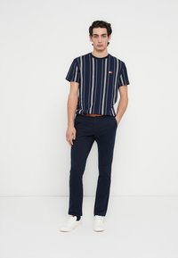 Μπλουζάκι με ναυτικές ρίγες σε μπλε χρώμα και λευκές γραμμές, κοντά μανίκια και μικρό λογότυπο, συνδυασμένο με σκούρο παντελόνι και λευκά αθλητικά παπούτσια. Άνετη εφαρμογή.