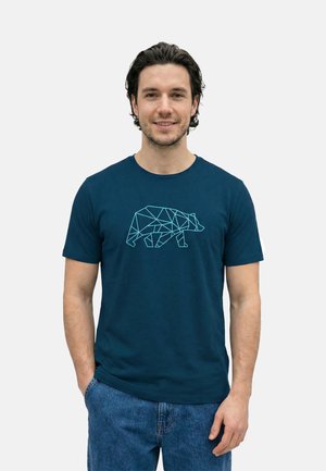 Mann mit kurzen dunklen Haaren, der ein dunkelblaues T-Shirt mit einem geometrischen Bärendesign trägt, lächelt, mit einer Hand in der Tasche und hellblauen Jeans.