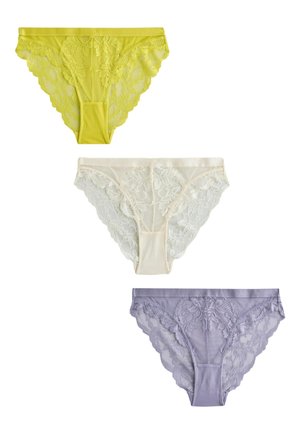 Kobieta w żółtym koronkowym biustonoszu i dopasowanej koronkowej bieliźnie, stojąca na tle gładkiej beżowej ściany.