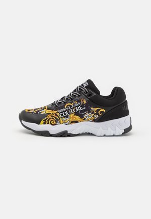 Baskets noir et blanc Versace Jeans Couture avec motif baroque jaune et texte de la marque sur le côté et la languette, montrées de profil.
