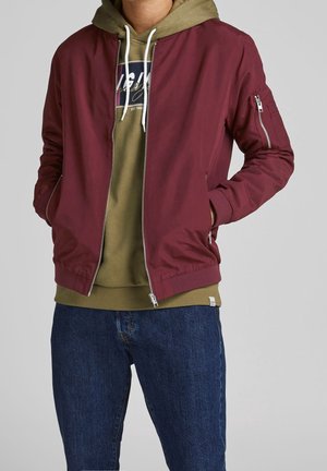 Mann trägt eine burgunderrote Bomberjacke über einem olivgrünen Hoodie mit weißen Kordeln und dunkelblauen Jeans, Hände in den Jackentaschen.