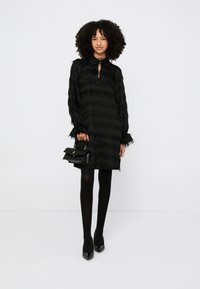 Γυναίκα με σγουρά μαλλιά που φοράει υφασμάτινο μαύρο φόρεμα, μαύρες καλσόν, μαύρες μυτερές γόβες και κρατά μια μικρή μαύρη τσάντα σε λευκό φόντο.