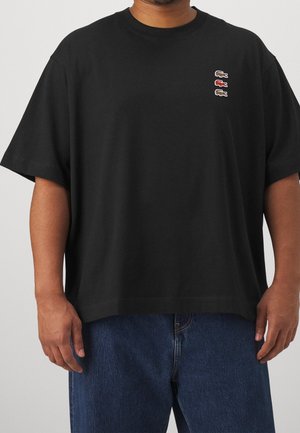 Musta puuvillainen t-paita, jossa on rento istuvuus. Paidassa on pyöreä pääntie, lyhyet hihat ja kolme pientä kirjailtua logoa rinnassa.