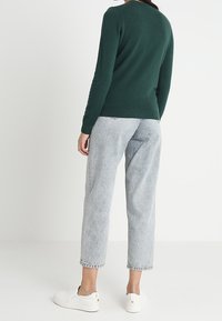 Mörkgrön stickad tröja, slim fit, rund halsringning, kombinerad med ljusgrå cropped jeans och vita slip-on skor. Mjuk textur, avslappnad stil.