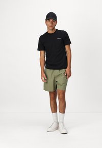 Camiseta negra con logo, pantalones cortos oliva claros, zapatillas blancas y calcetines blancos. El conjunto presenta un ajuste casual y una tela suave.