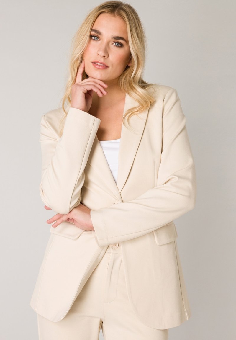Beige op maat gemaakte blazer met notch revers, zijzakken en een enkele knoopsluiting, gecombineerd met een witte top. Zachte stoftextuur.