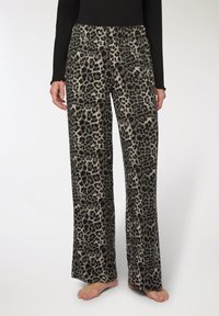 Persona che indossa pantaloni a gamba larga con stampa leopardata e una maglietta nera a maniche lunghe, in piedi a piedi nudi contro uno sfondo semplice.