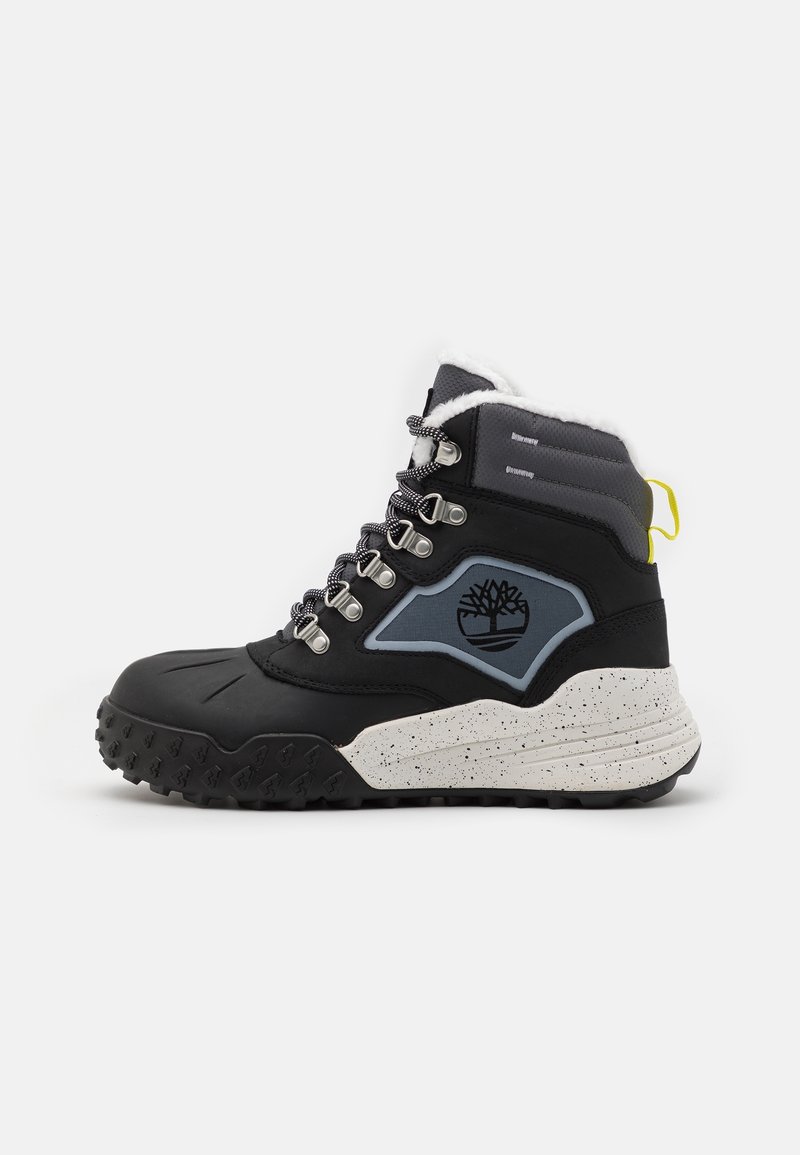 Botte mi-haute noire avec des accents gris, semelle blanche mouchetée, œillets en métal et col en fourrure. Présente un logo sur le côté et un profil de semelle texturé.