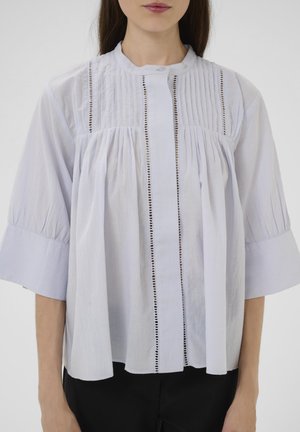 White blouse with a high collar, vertical pleats, and eyelet accents, featuring wide sleeves and a loose, flowing silhouette.