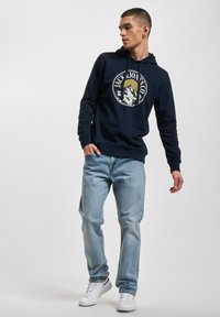 Navy Kapuzenpullover mit Logo-Druck, der ein Berg- und Sonnenmotiv zeigt, kombiniert mit hellblauen Jeans und weißen Sneakers.