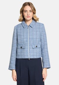 Femme portant une veste zippée texturée bleu et blanc avec poches à rabat, associée à un pantalon large bleu marine, debout devant un fond uni.