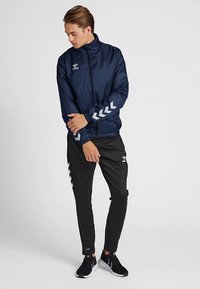 Marineblå vindjakke med lynlås, reflekterende accenter på ærmerne, kombineret med sorte, indsnævrede bukser med hvidt chevronmønster.