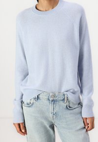 Suéter de cachemira azul claro con cuello redondo, mangas raglán y puños acanalados, combinado con jeans de mezclilla azul claro, mostrando un corte relajado.