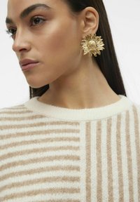 Goldene, sonnenblumenförmige Ohrringe neben einer Person, die einen cremigen Pullover mit abwechselnden beigen Streifen trägt. Glatte Textur, natürliche Designelemente.