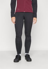 Fekete, elasztikus leggings, amely fitted sziluettel rendelkezik. Egy bordó, zipzáros dzsekivel van párosítva, fekete ujjakkal és fényvisszaverő logó részletekkel.