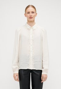 Blouse crème à manches longues, en tissu texturé, avec des bordures en dentelle le long de la patte de boutonnage et du col. Portée avec un pantalon noir.