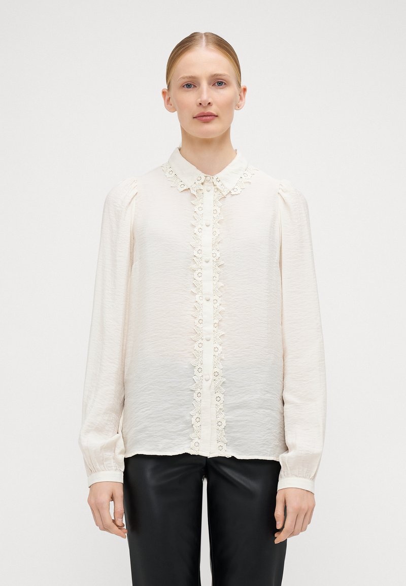 Blouse crème à manches longues, en tissu texturé, avec des bordures en dentelle le long de la patte de boutonnage et du col. Portée avec un pantalon noir.
