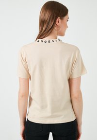 Camiseta de manga corta color beige en tejido suave, con un texto negro "ANGESTR." en el cuello. Diseño sencillo con un corte relajado.