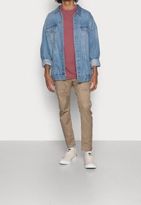 Lichtblauwe spijkerjacket, in combinatie met een bordeauxrode t-shirt, tan cargobroeken en beige sneakers. Casual outfit met opgerolde mouwen.