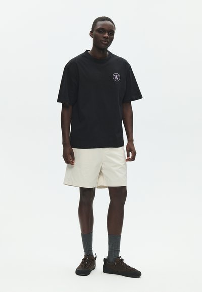 Νέος άνδρας όρθιος, φοράει μαύρο oversized T-shirt με λογότυπο, μπεζ σορτς, γκρι κάλτσες και καφέ αθλητικά παπούτσια σε απλό φωτεινό φόντο.