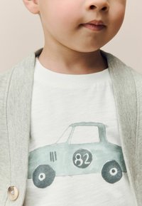 Lichtgrijze cardigan over een wit shirt met korte mouwen, met een pastelgroene autografiek met het nummer 82, zwarte wielen en eenvoudige omranding.