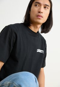 Czarna bawełniana koszulka z krótkim rękawem i okrągłym dekoltem, z białym logotypem "Carhartt" po lewej stronie klatki piersiowej.