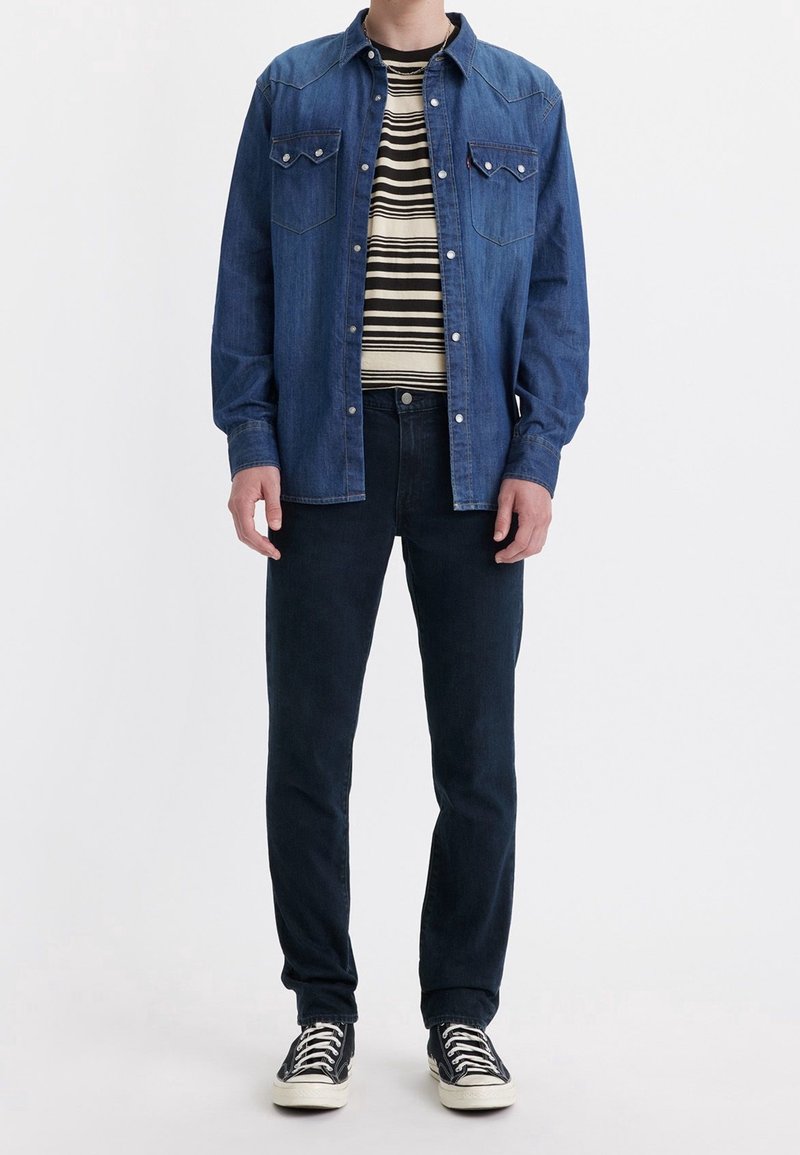 Homme portant une veste en jean bleue sur une chemise rayée noire et blanche, un jean foncé et des baskets basses noires, debout devant un fond blanc.