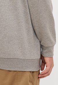 Sweatshirt gris avec un col et un ourlet côtelés, texture douce et coupe décontractée. Main reposant sur la taille, associé à un pantalon beige.