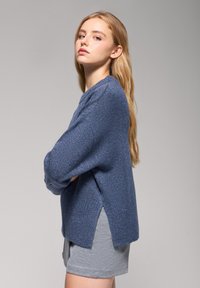 Niebieski sweter dzianinowy o prążkowanej teksturze, z obniżonymi ramionami i bocznymi rozcięciami. Stylizowany z szarymi szortami, na neutralnym tle.