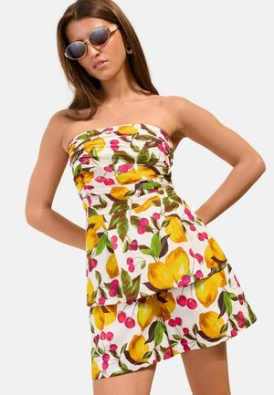 Frau trägt trägerloses weißes Kleid und Shorts mit gelbem Zitronen- und rotem Kirschmuster, posiert mit Händen in der Hüfte und Sonnenbrille.