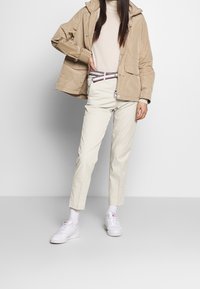 Giacca impermeabile beige con cappuccio, indossata sopra un maglione leggero, abbinata a pantaloni color crema e sneakers bianche. Design minimal, vestibilità casual.