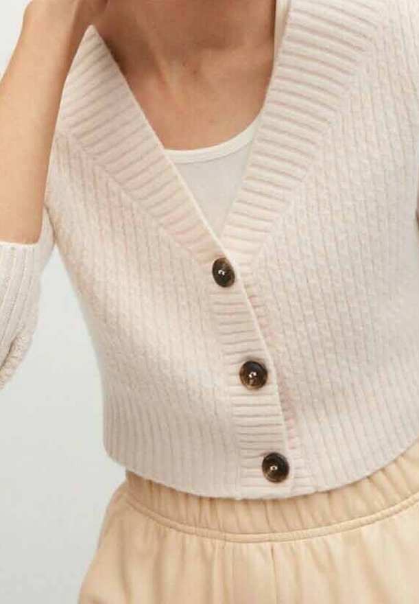 Femme portant un cardigan court côtelé crème avec trois boutons marron sur un débardeur blanc et un pantalon beige taille haute.