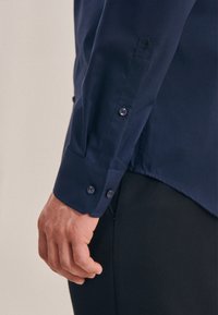 Marineblauwe lange mouwen shirt met een knoop manchet met twee knopen en een subtiele geborduurde detail op de mouw. Soepele, gepolijste stoftextuur.