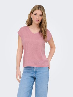 Femme aux longs cheveux blonds portant un haut rose à manches courtes et un jean bleu clair, debout avec une main sur la hanche devant un fond uni.