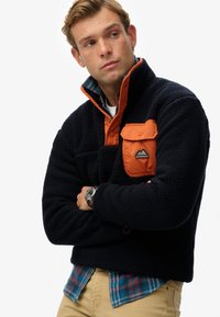 Dunkelblauer Fleece-Pullover mit orangefarbener Druckknopfkrause und Brusttasche, der eine strukturierte Oberfläche sowie ein Logo-Patch auf der Tasche aufweist.