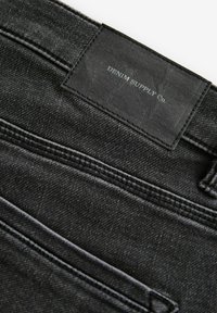 Material din denim negru cu un finisaj texturat, având o etichetă cu inscripția "DENIM SUPPLY Co." împreună cu detalii de coasere și conturul unui buzunar.