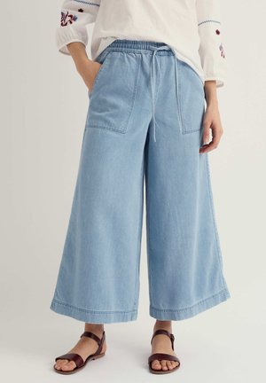 Nő világoskék, széles szárú, rövidített farmernadrágot visel elülső zsebekkel, fehér hímzett blúzzal és barna bokapántos szandállal.