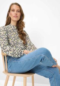 Let bluse med mønster, høj hals og lange ærmer. Kombineret med lyseblå denimjeans. Model siddende på en træstol.