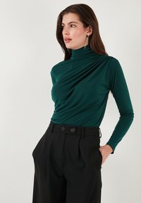 Vrouw met lang bruin haar, gekleed in een groene gerimpelde coltrui en hoge zwarte broeken, staand met handen in haar zakken.