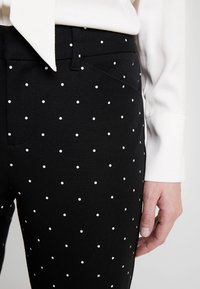 Czarne spodnie materiałowe w białe kropki, o slim fit, z kieszeniami z przodu i teksturowanej powierzchni. Ręka umieszczona w pobliżu pasa.