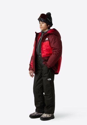 Chaqueta aislante roja y negra, capa base gris, pantalones de nieve negros, botas de camuflaje y gorro negro con gafas.