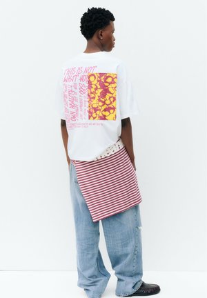 Jeune personne portant un t-shirt blanc graphique avec un design rose et jaune, un tissu rayé rentré à l'arrière, un jean bleu ample et des chaussures noires.