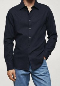 Chemise à manches longues bleu marine en tissu texturé avec patte de boutonnage, col pointu et coupe décontractée, portée avec un jean bleu.