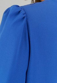Blaue Bluse mit Puffärmeln, aus glattem Stoff gefertigt. Verfügt über zarte Falten an der Schulter und ein sauberes, minimalistisches Design.