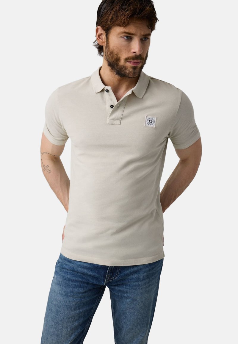 Homme barbu portant un polo beige à manches courtes avec un patch logo sur la poitrine et un jean bleu, debout, mains derrière le dos, regardant vers le bas et à droite.