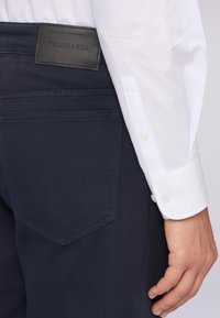 Pantalones navy hechos de una tela tejida con una textura suave, que presentan una etiqueta de cuero negro y una manga de camisa blanca en el encuadre.