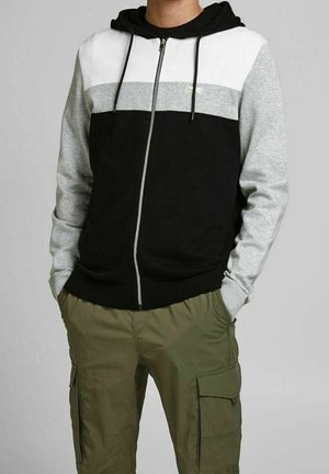 Personne portant un sweat à capuche zippé avec blocs de couleurs noirs, gris et blanc, associé à un pantalon cargo vert olive, les mains dans les poches.