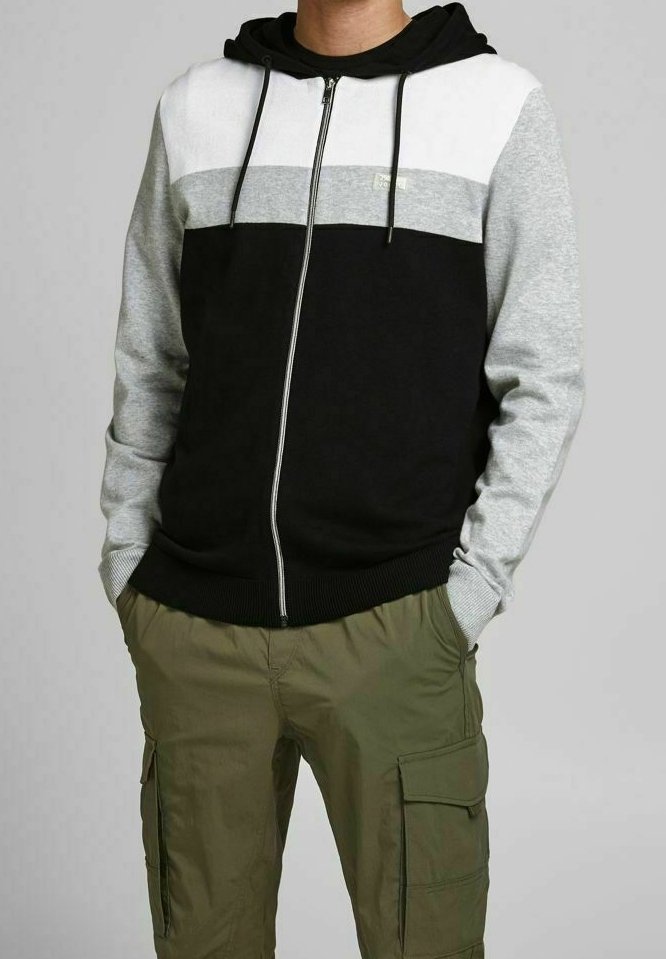 Personne portant un sweat à capuche zippé avec blocs de couleurs noirs, gris et blanc, associé à un pantalon cargo vert olive, les mains dans les poches.