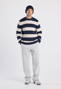 Giovane uomo che indossa un berretto blu navy, un maglione a righe blu e bianche, pantaloni felpati grigi chiari e sneakers bianche, in piedi con le mani in tasca.